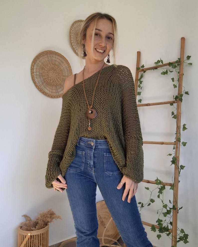 May include: An olive green, loose-knit, long-sleeved sweater with a wide neckline, paired with blue denim jeans featuring front patch pockets. A long brown leather cord necklace with a circular wooden pendant hangs around the neck.