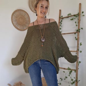 May include: An olive green, loose-fitting, open-knit sweater with a wide boat neckline and long, dropped-shoulder sleeves. The garment features a textured, airy stitch pattern and is paired with blue denim jeans, creating a casual, layered look.