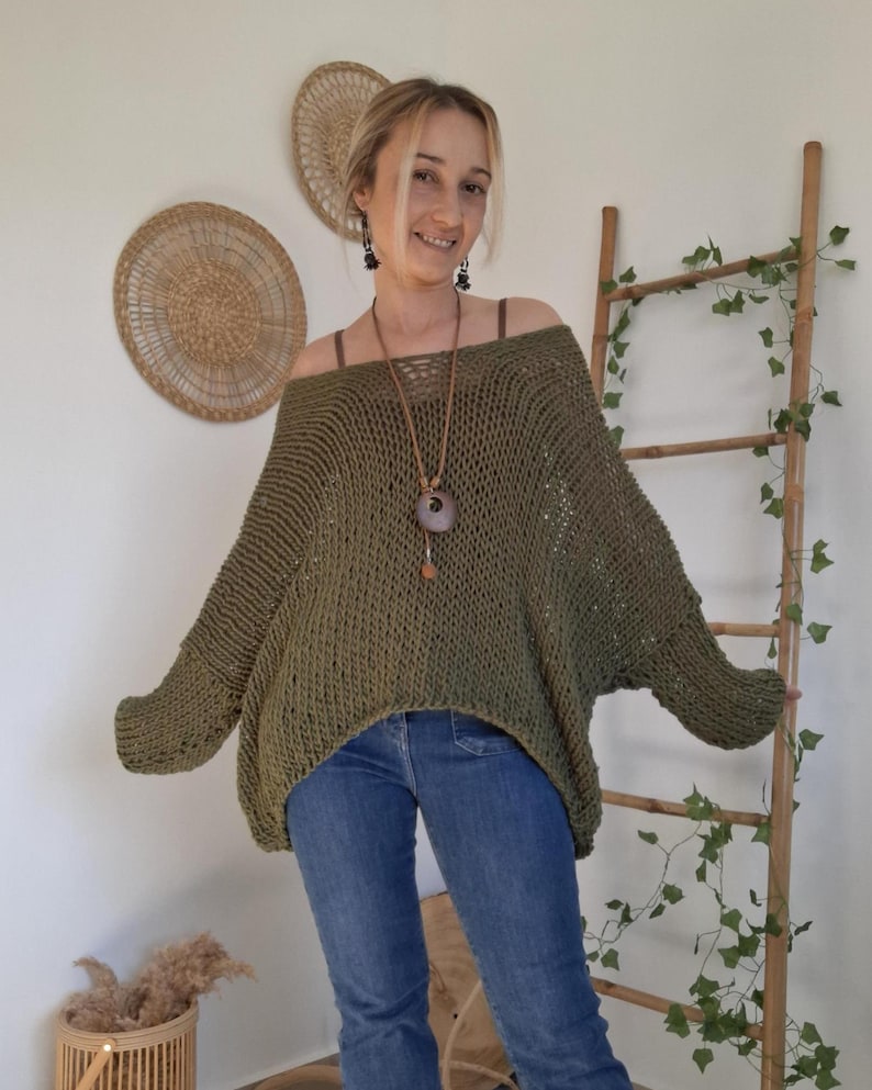 May include: An olive green, loose-fitting, open-knit sweater with a wide boat neckline and long, dropped-shoulder sleeves. The garment features a textured, airy stitch pattern and is paired with blue denim jeans, creating a casual, layered look.