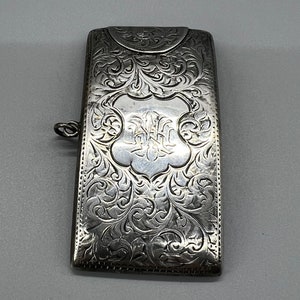 May include: A silver match safe with an ornate engraved design. The front features a floral pattern with a central shield.