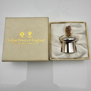 Könnte beinhalten: Ein silberner Flaschenverschluss mit einer dekorativen goldfarbenen Spitze, präsentiert in einer cremefarbenen Schachtel. Die Schachtel ist mit "Arthur Price of England" mit goldenem Emblem und Text beschriftet. Der Verschluss wird auf weißem Satin-ähnlichem Stoff präsentiert.