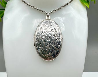Puede incluir: Collar con colgante de medallón ovalado plateado con intrincado grabado floral. El medallón cuelga de una delicada cadena y se exhibe sobre un busto blanco. La superficie del medallón es muy detallada, mostrando un diseño clásico y vintage.