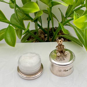 Puede incluir: Un recipiente plateado con un pequeño oso de peluche dorado encima, junto a un recipiente con borde plateado con una bola de algodón blanca en su interior. El recipiente con el oso tiene texto grabado.