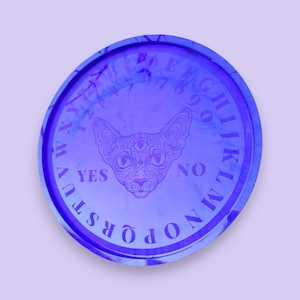 May include: A purple and blue circular ouija board with a cat design in the center. The board has the letters of the alphabet, numbers 0-9, and the words "yes" and "no" printed on it.