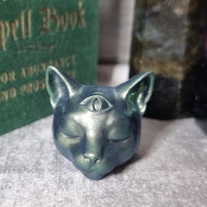Op de afbeelding: Een metallic teal kattenhoofdsculptuur met een derde oog op het voorhoofd. De ogen van de kat zijn gesloten en de sculptuur heeft een gladde, iriserende afwerking. De achtergrond bevat een groen boek met gouden letters.