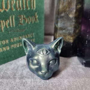 Op de afbeelding: Een metallic teal kattenhoofdsculptuur met een derde oog. De sculptuur staat voor een groen boek met gouden letters met de tekst "Wealth Spell Book". De sculptuur heeft een glad, reflecterend oppervlak. De kattenkop is ongeveer 5 cm hoog.