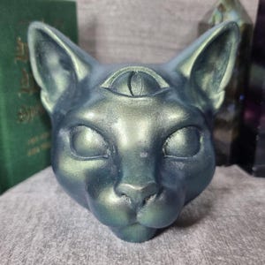 Op de afbeelding: Een teal-kleurige kattenhoofdsculptuur met een derde oog op het voorhoofd. De sculptuur heeft een metallic glans en gedetailleerde gelaatstrekken, waaronder grote, gesloten ogen en een kleine neus. Decoratief item.