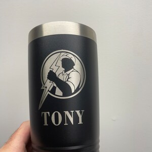 Op de afbeelding: Een zwarte roestvrijstalen beker met een zilveren deksel. De beker heeft een zilveren gegraveerd beeld van een elektricien die een bliksem vasthoudt en de naam "TONY" onder het beeld.