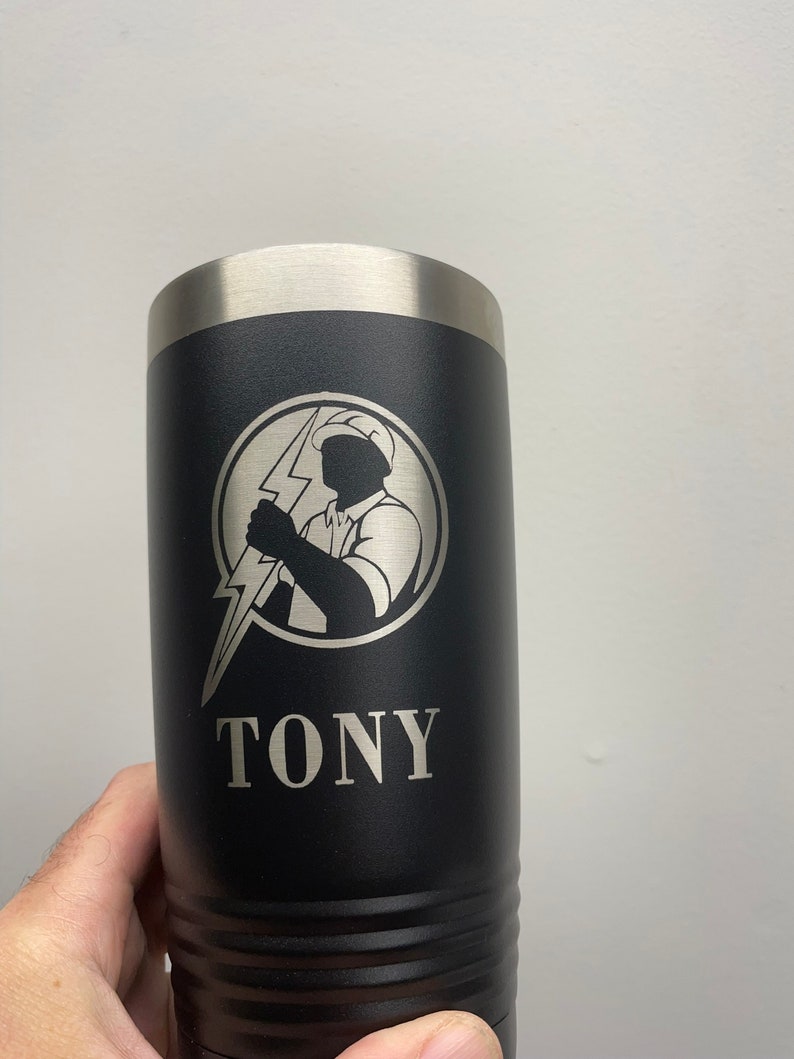 Op de afbeelding: Een zwarte roestvrijstalen beker met een zilveren deksel. De beker heeft een zilveren gegraveerd beeld van een elektricien die een bliksem vasthoudt en de naam "TONY" onder het beeld.