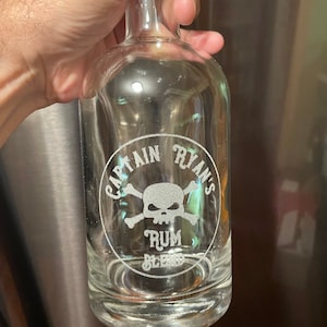 May include: A clear glass bottle with a skull and crossbones design etched on the front. The bottle says "Captain Ryan's Rum Blend".