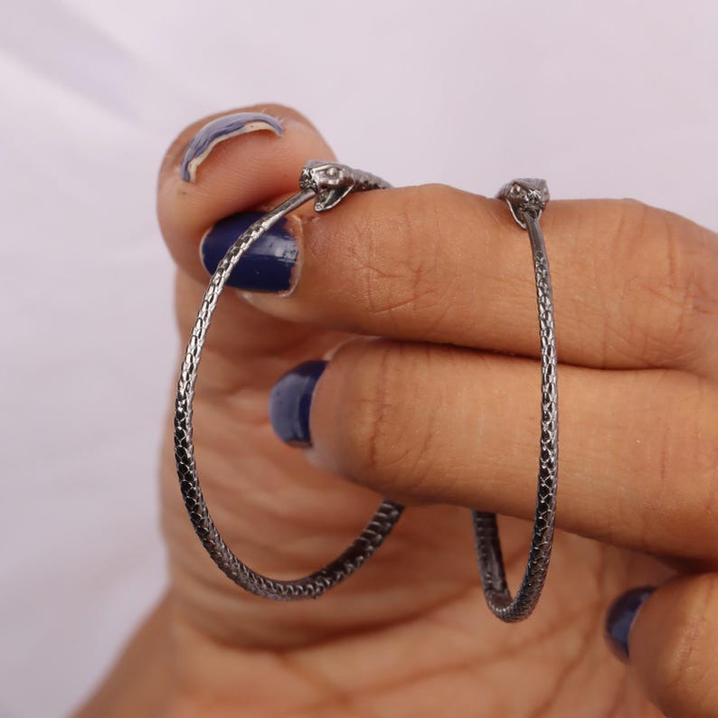 May include: A pair of silver hoop earrings with snake head accents. The earrings are textured and have a slightly oxidized finish.