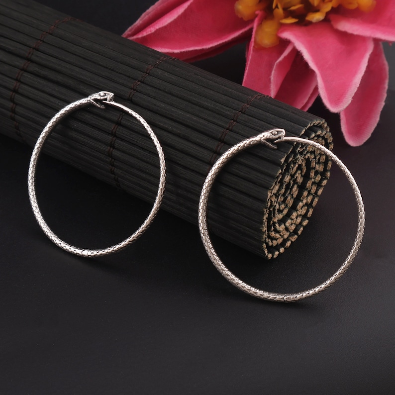 May include: A pair of silver hoop earrings with a snake head design. The earrings are on a black background.