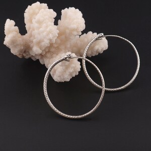 May include: A pair of silver snake hoop earrings. The earrings are made of a textured metal that resembles snake scales. The earrings are hanging from a white coral-like surface.