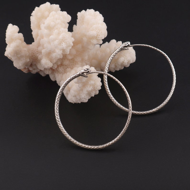 May include: A pair of silver snake hoop earrings. The earrings are made of a textured metal that resembles snake scales. The earrings are hanging from a white coral-like surface.
