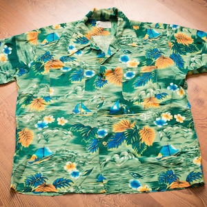 May include: Green short-sleeve button-down shirt with a tropical print. The shirt features a pattern of sailboats, palm trees, and flowers in shades of blue, yellow, and orange. The shirt is laid flat on a wooden surface.