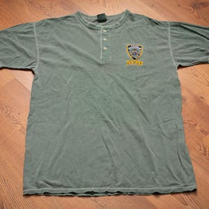 May include: A sage green short-sleeve henley shirt featuring a three-button placket at the neckline. The left chest displays an embroidered NYPD police department shield emblem in blue, white, and gold, with the text NYPD printed in yellow below.