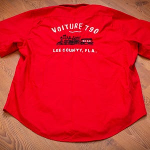 May include: A bright red short-sleeved button-down shirt featuring a graphic on the back. The print includes the text "VOITURE 790" above a black illustration of a train car, with the text "LEE COUNTY, FLA." printed in white below the graphic.