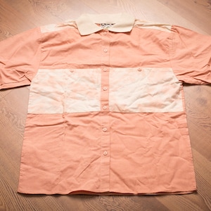 May include: A short-sleeved button-down shirt in a peach color with a cream-colored collar and chest panel. The shirt has a color-blocked design with a cream-colored section across the chest and a cream-colored collar.