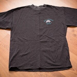 May include: A dark gray t-shirt with a ribbed texture. The shirt has a small embroidered design on the front that says "San Antonio Texas" with a small armadillo graphic.