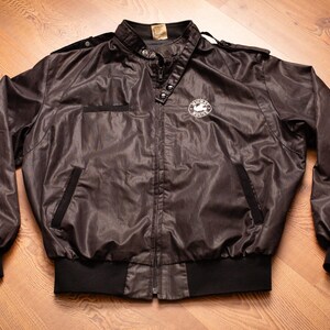 May include: A black nylon bomber jacket with a zipper closure and a white logo on the left chest. The jacket has a ribbed collar, cuffs, and waistband.