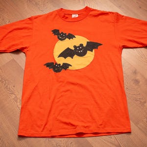 May include: A bright orange short-sleeved t-shirt featuring a large yellow circular patch in the center. Three black bat appliques are stitched onto the yellow circle, each with small white fangs and eyes, creating a layered graphic design.