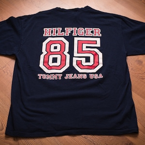 May include: A navy blue t-shirt with a white and red graphic print of the text "HILFIGER" and the number "85". The text "TOMMY JEANS USA" is printed below the number.