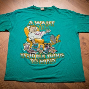 May include: A green t-shirt with a cartoon character sitting in a chair and eating junk food. The text on the shirt reads "A waist is a terrible thing to mind".
