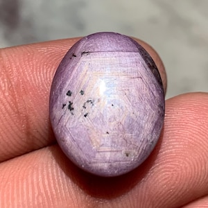 May include: Oval-shaped, lavender-colored gemstone with a star-like pattern. The stone has a polished surface with a subtle sheen, and small dark inclusions are visible. The gemstone is held between fingers.