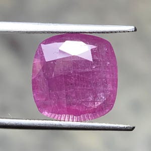 May include: A cushion-cut pink gemstone with a faceted surface and visible internal grid-like inclusions. The translucent stone displays a vibrant magenta hue and is held between the two metal tips of a pair of tweezers for display.