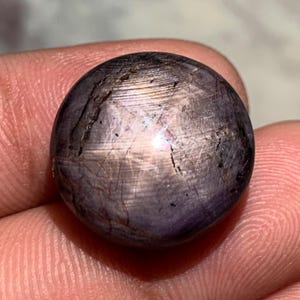 May include: A round, cabochon-cut star sapphire gemstone with a deep purplish-gray hue. The surface displays a distinct six-rayed star effect, known as asterism, caused by light reflecting off internal needle-like inclusions within the stone.