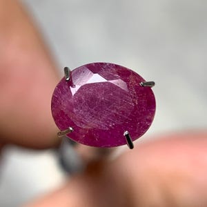 Può includere: Una gemma di rubino sfaccettata, di forma ovale, tenuta da quattro griffe d'argento. Il rubino è di un colore rosso intenso e ricco, con inclusioni interne visibili. L'incastonatura è semplice, permettendo alla gemma di essere il punto focale.