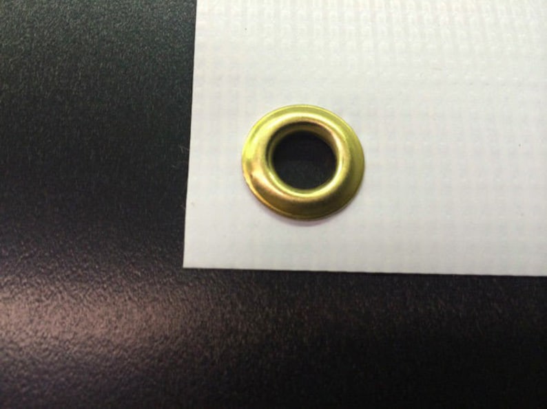 May include: A close-up shot of a gold-colored grommet, a metal ring used to reinforce holes in fabric or other materials. The grommet is set against a white background, with a dark surface below.