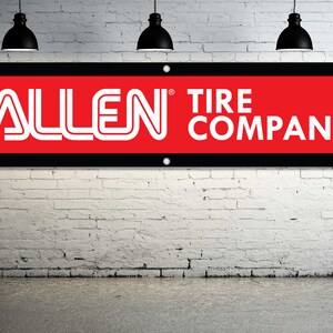 May include: A rectangular banner with the text "ALLEN TIRE COMPANY" in white lettering. The banner has a red background with a black border and is mounted on a white brick wall. Three black pendant lights hang above.
