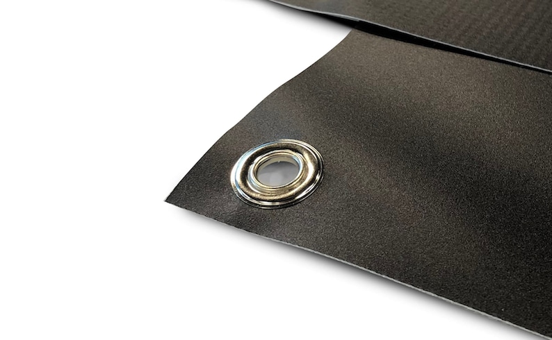 May include: Close-up of a section of black material with a silver grommet. The material has a textured surface and is folded over, revealing the grommet's shiny metal finish. The background is white.