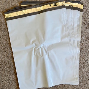 May include: A stack of white poly mailer shipping envelopes with a gold and black border. The envelopes are made of a durable, waterproof material and are designed for shipping various items. The envelopes are arranged on a brown carpet.