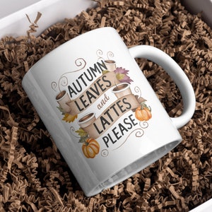 May include: White ceramic mug with the text "AUTUMN LEAVES and LATTES PLEASE" in a banner design, with coffee cups, pumpkins, and fall leaves. The mug is nestled in brown paper filler inside a white box.