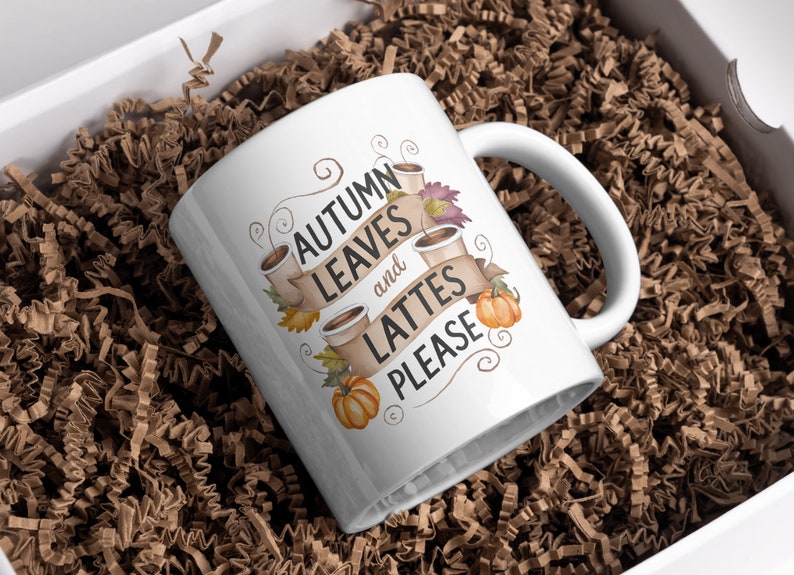 May include: White ceramic mug with the text "AUTUMN LEAVES and LATTES PLEASE" in a banner design, with coffee cups, pumpkins, and fall leaves. The mug is nestled in brown paper filler inside a white box.