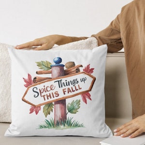 May include: White square pillow with a watercolor design featuring a signpost that reads "Spice Things up THIS FALL". The signpost is decorated with cinnamon sticks, leaves, and a nutmeg. The pillow is on a white surface.