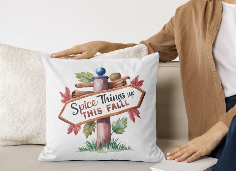 May include: White square pillow with a watercolor design featuring a signpost that reads "Spice Things up THIS FALL". The signpost is decorated with cinnamon sticks, leaves, and a nutmeg. The pillow is on a white surface.
