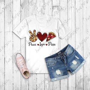 May include: White t-shirt with a peace sign, heart, and pizza slice graphic. The graphic is red, yellow, and brown. The text "Peace Love Pizza" is printed below the graphic. The t-shirt is on a wooden surface with a pair of blue denim shorts and pink sneakers.
