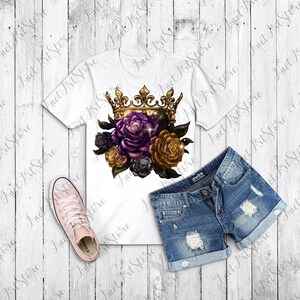 Op de afbeelding: Een wit T-shirt met een gouden kroon en paarse, gouden en zwarte rozen bedrukt op de voorkant. Het T-shirt ligt op een houten oppervlak met een spijkerbroek en roze sneakers.