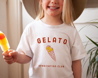 May include: A white t-shirt with the text "GELATO APPRECIATION CLUB" in brown lettering. A cartoon image of a gelato cone with three scoops of yellow gelato is centered on the shirt.