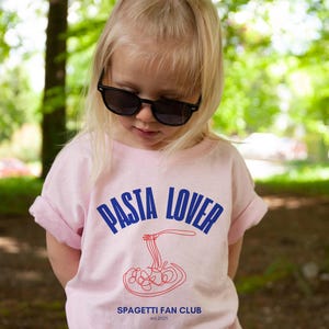 May include: A young person wearing a pink t-shirt with the text "PASTA LOVER" in blue and a red spaghetti graphic. The text "SPAGHETTI FAN CLUB est. 2023" is printed below the graphic.