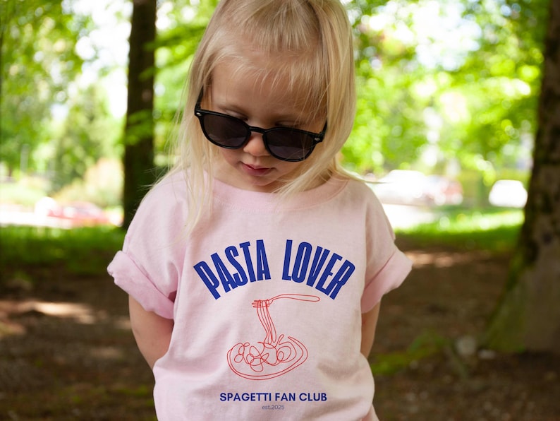 May include: A young person wearing a pink t-shirt with the text "PASTA LOVER" in blue and a red spaghetti graphic. The text "SPAGHETTI FAN CLUB est. 2023" is printed below the graphic.