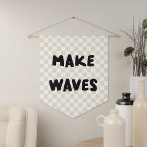 Puede incluir: Una pancarta de tela a cuadros beige y blanca con el texto "MAKE WAVES" en negro. La pancarta está colgada de una varilla de madera con cuerda.