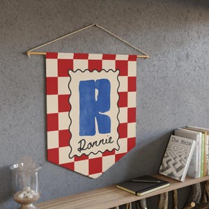 Op de afbeelding: Een decoratieve wandbanner met een rood-wit geblokt patroon. De banner toont de blauwe letter "R" en de naam "Ronnie". De banner is opgehangen aan een houten stok met een natuurlijk touw. Geschikt voor een kinderkamer.