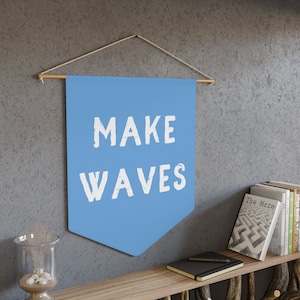 Puede incluir: Una pancarta azul claro con el texto "MAKE WAVES" en letras blancas. La pancarta está colgada de una varilla de madera con cuerda.