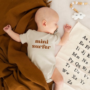 Puede incluir: Un bebé con un mono beige con el texto "mini surfer" en letras marrones. El bebé está tumbado en una manta blanca con una manta marrón debajo. Hay una nube blanca con ojos negros y un collar de cuentas de madera en la manta.