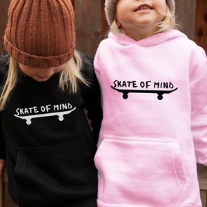 May include: Two children wearing hoodies with the text "Skate of Mind" and a graphic of a skateboard. One hoodie is black with white text and the other is pink with black text.