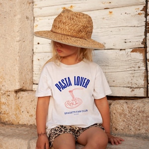 May include: A young person wearing a white t-shirt with the text "PASTA LOVER" and "SPAGHETTI FAN CLUB" in blue. The person is wearing a straw hat and has a brown and white animal print shorts on.